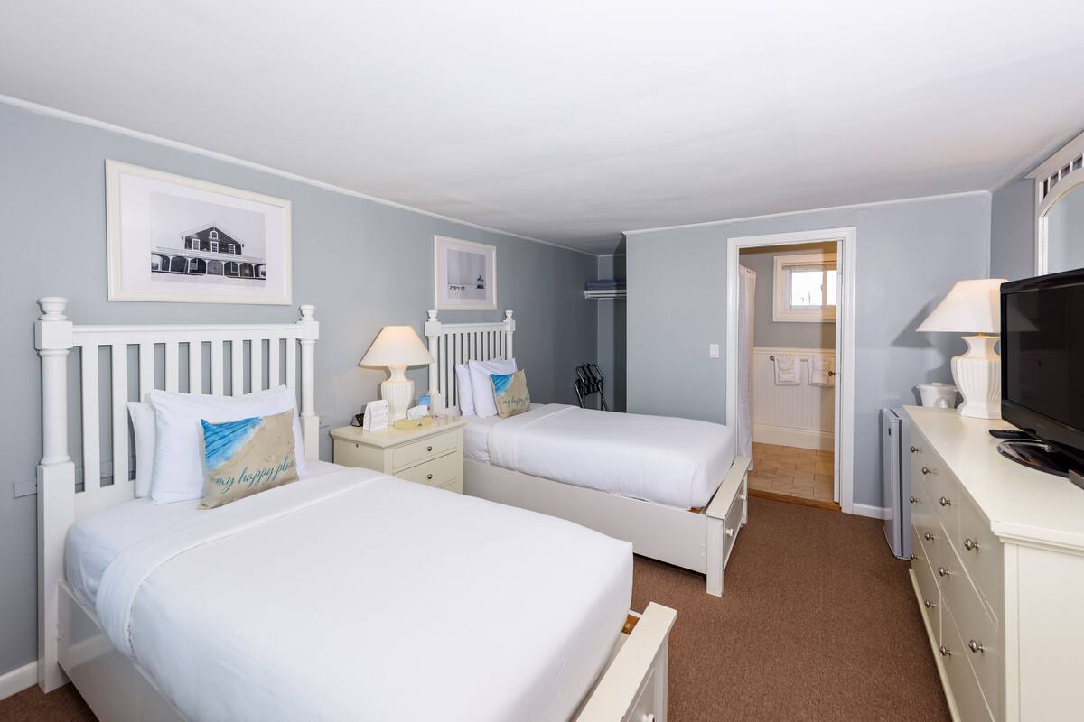 A bedroom with two single beds, white headboards, bedside tables with lamps, a dresser, a television, and framed pictures on the wall.
