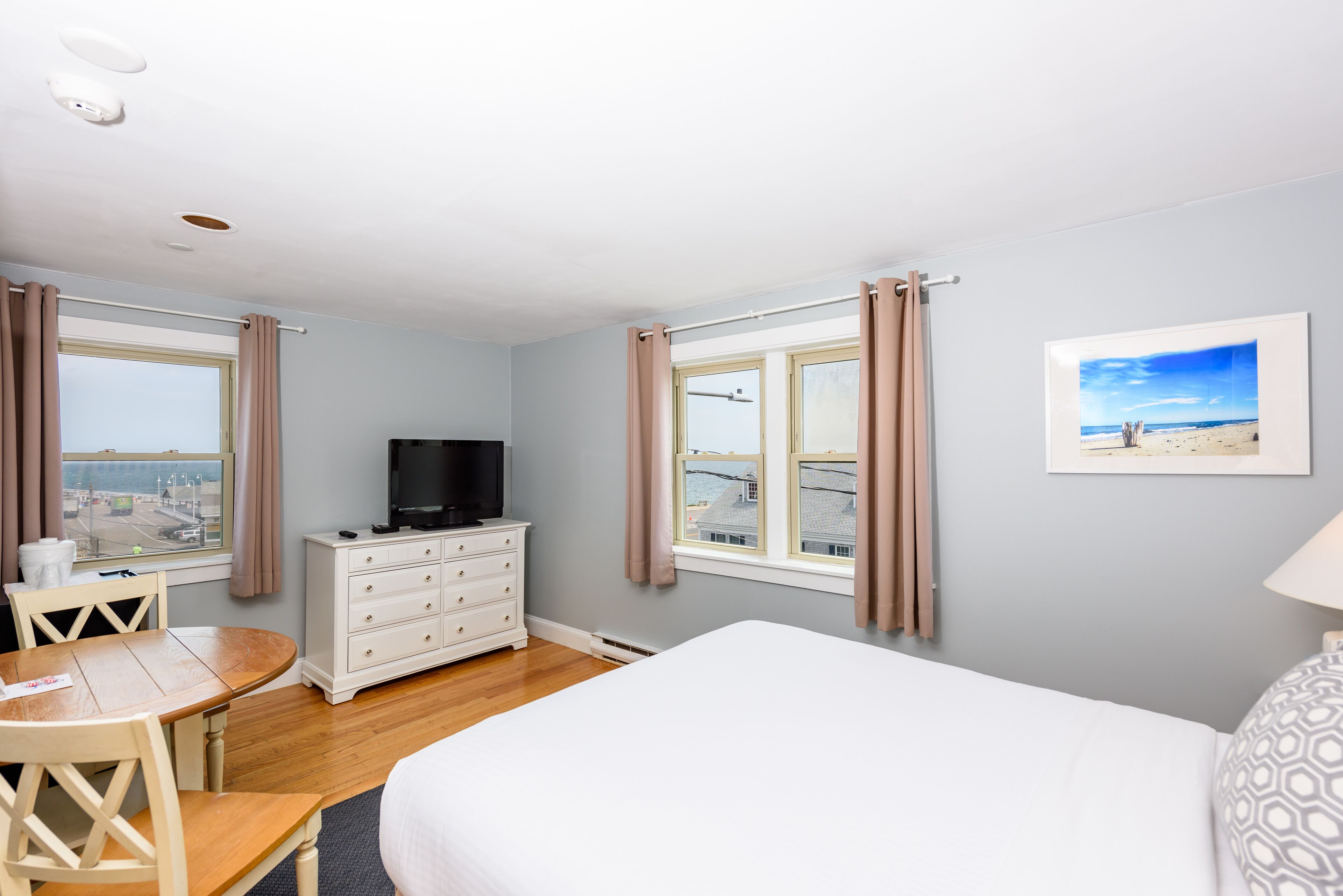 A hotel room with a bed, a TV on a dresser, a dining table, and two windows with curtains.
