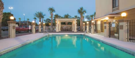 A swimming pool with a covered seating area, surrounded by palm trees and a building with a beige facade.