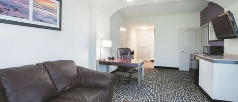 A modern hotel room with a leather sofa, a dining table, a kitchen area, and a framed picture on the wall.