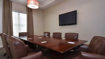 A conference room with a large wooden table, six brown leather chairs, a flat-screen TV mounted on the wall, and a window with curtains.
