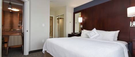 A hotel room with a large bed, a wooden headboard, a bedside table with a lamp, a mirror, and a bathroom visible through an open door.