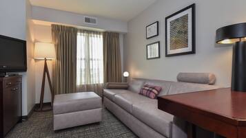A hotel room with a sofa, ottoman, a TV, a lamp, and framed pictures on the wall.