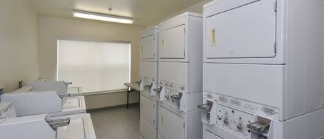 A laundry room with multiple stacked washers and dryers, a window with blinds, and a fluorescent ceiling light.