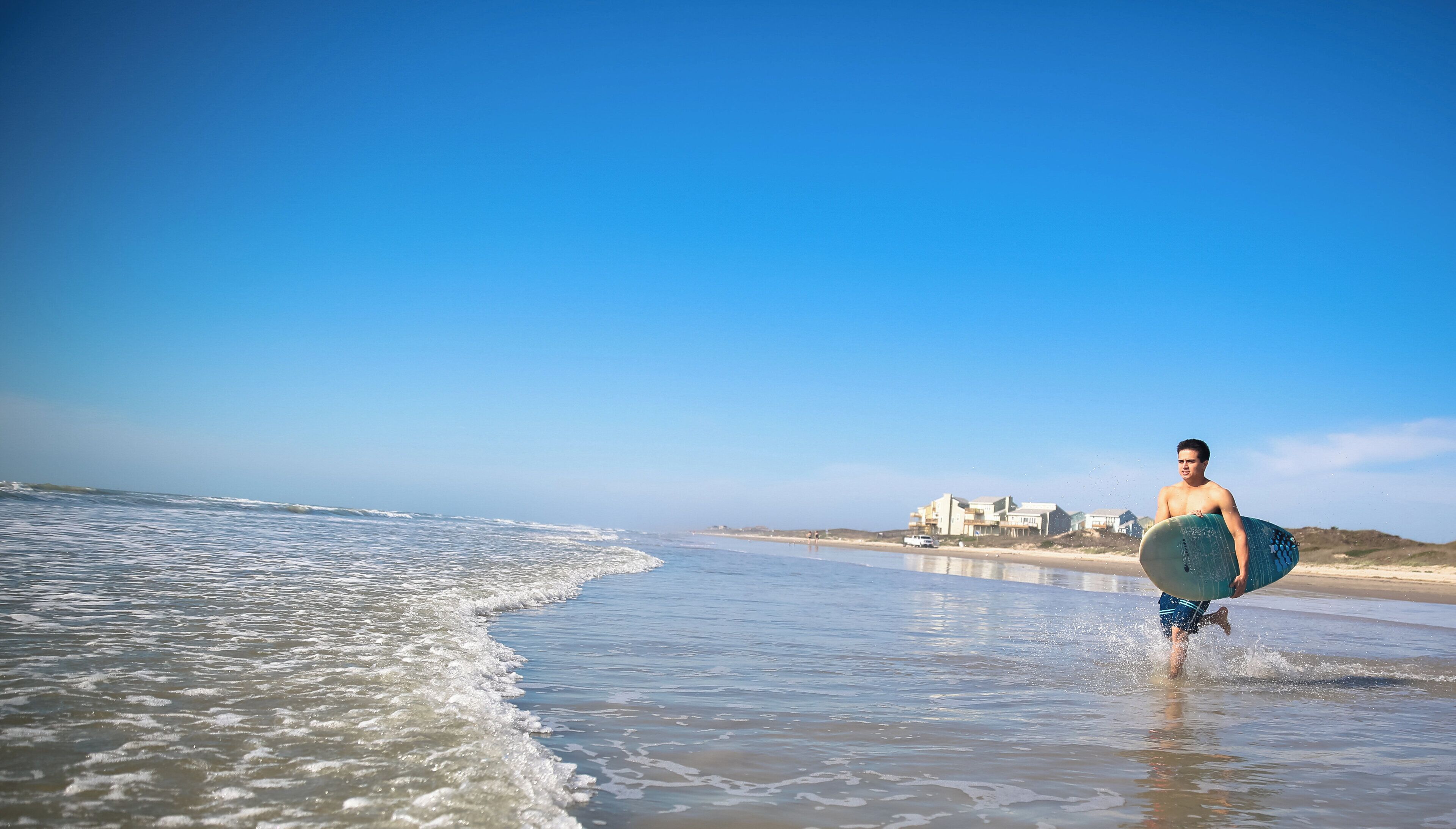 Eine Strandansicht mit einer Person, die einen Surfboard trägt, Wellen und Gebäuden im Hintergrund.