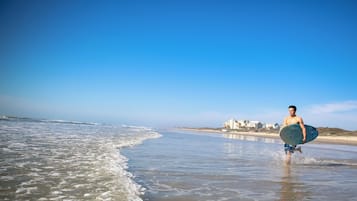 Eine Strandansicht mit einer Person, die einen Surfboard trägt, Wellen und Gebäuden im Hintergrund.