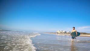 A beach scene with a person carrying a surfboard, waves, and buildings in the background. - Staybridge Suites Corpus Christi by IHG (Corpus Christi)
