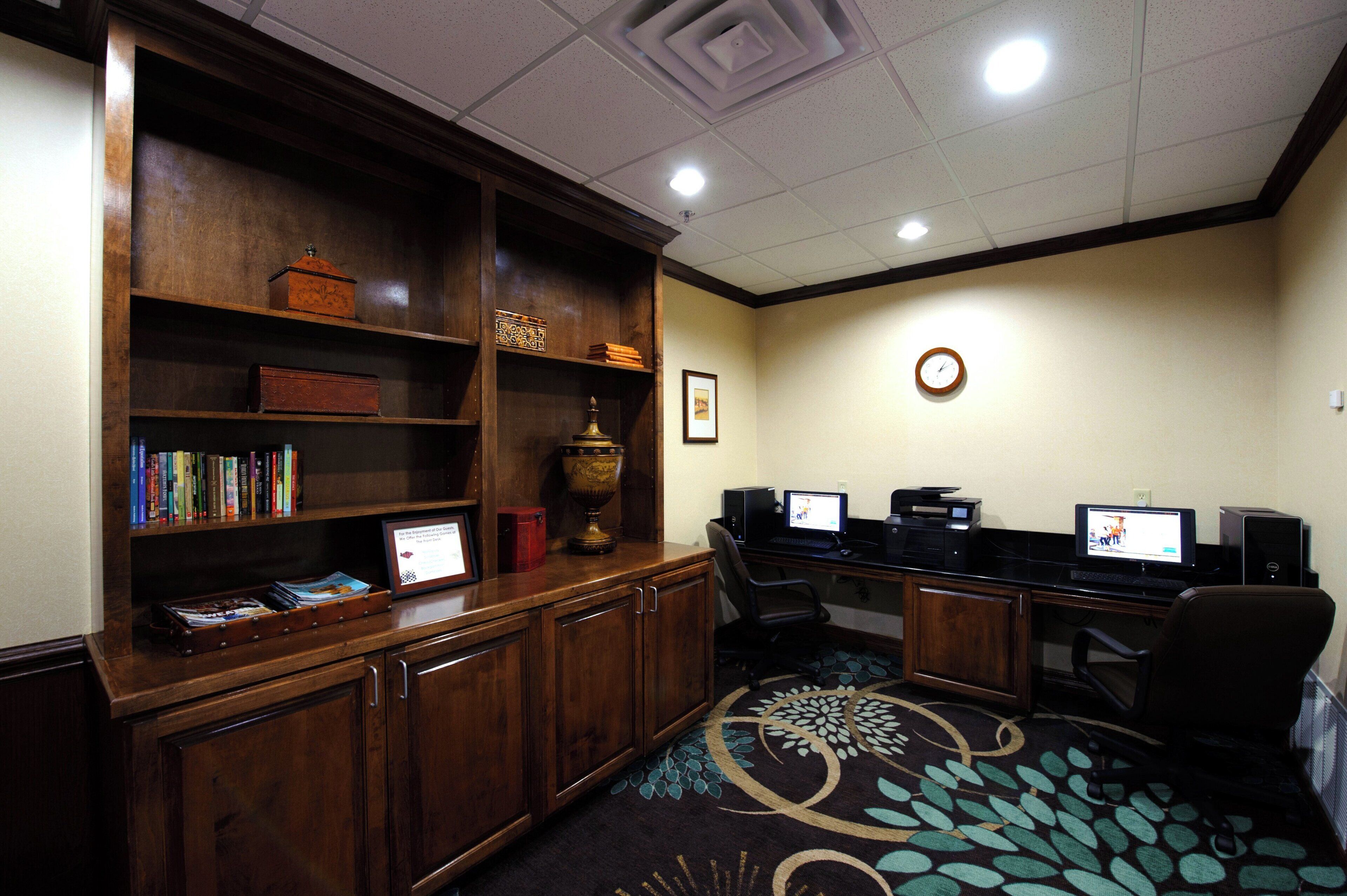 A room with a wooden desk, two computer monitors, a bookshelf, and a patterned carpet.