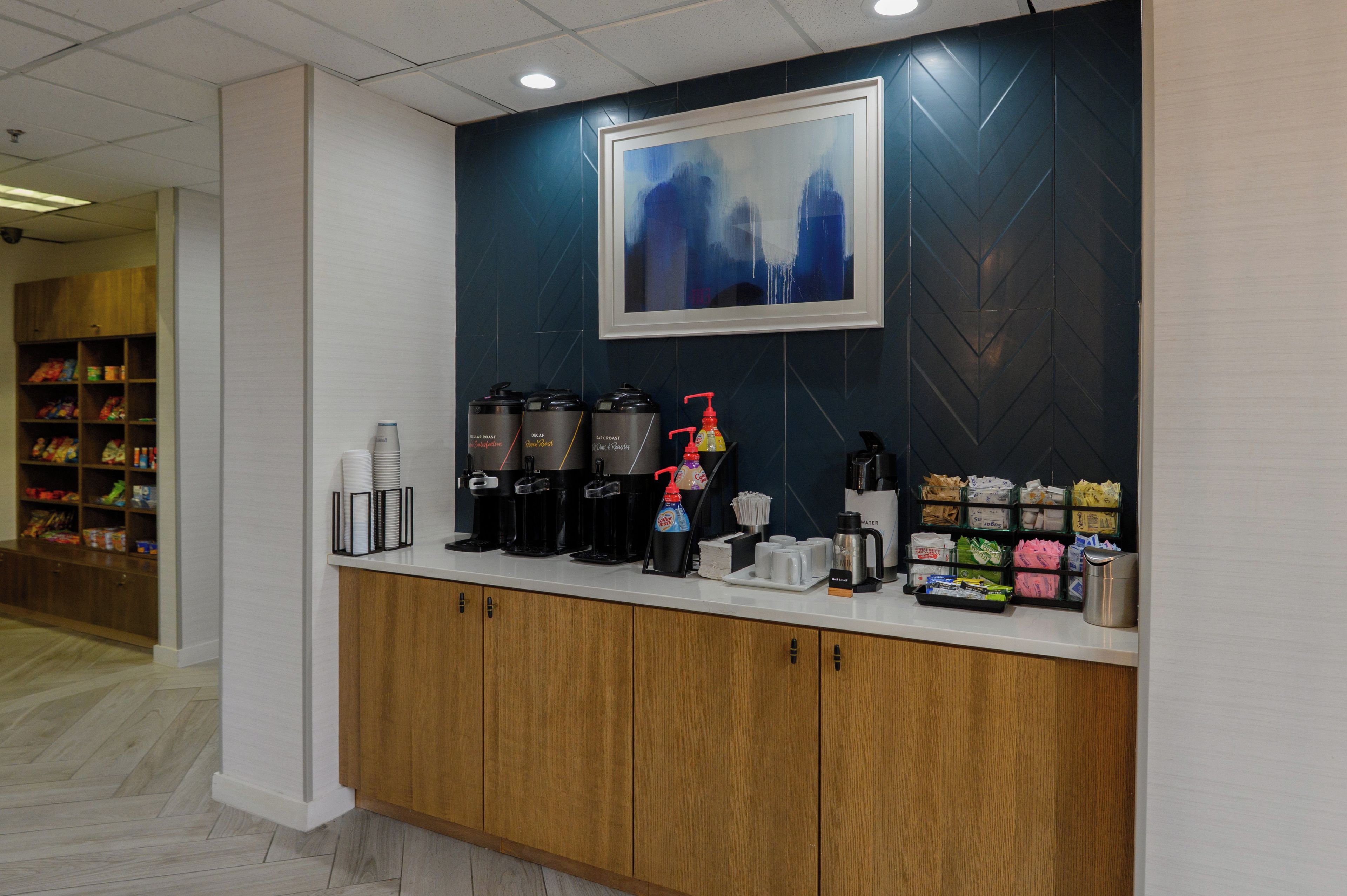 Zona de recepción con estación de café, dispensador de agua y un cuadro abstracto enmarcado en la pared.