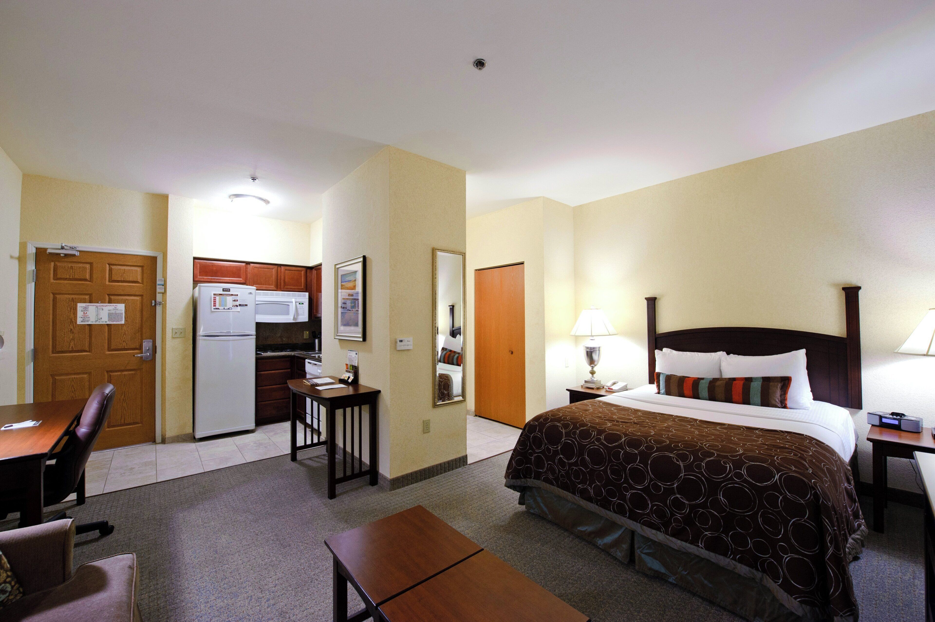 Una habitación de hotel con cama, escritorio, silla, kitchenette y una puerta.