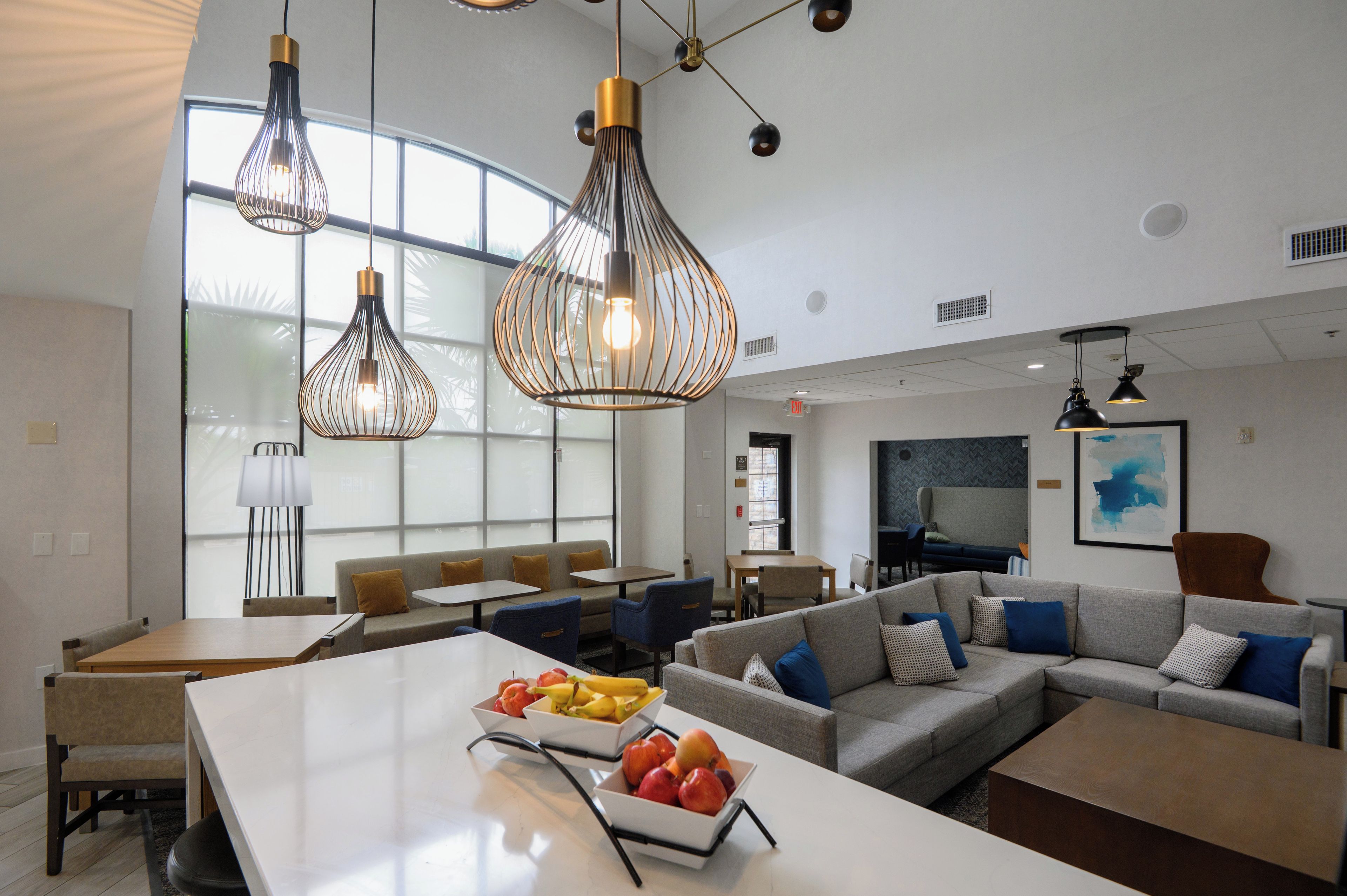 A modern hotel lobby with seating areas, a fruit bowl on a table, and pendant lighting.