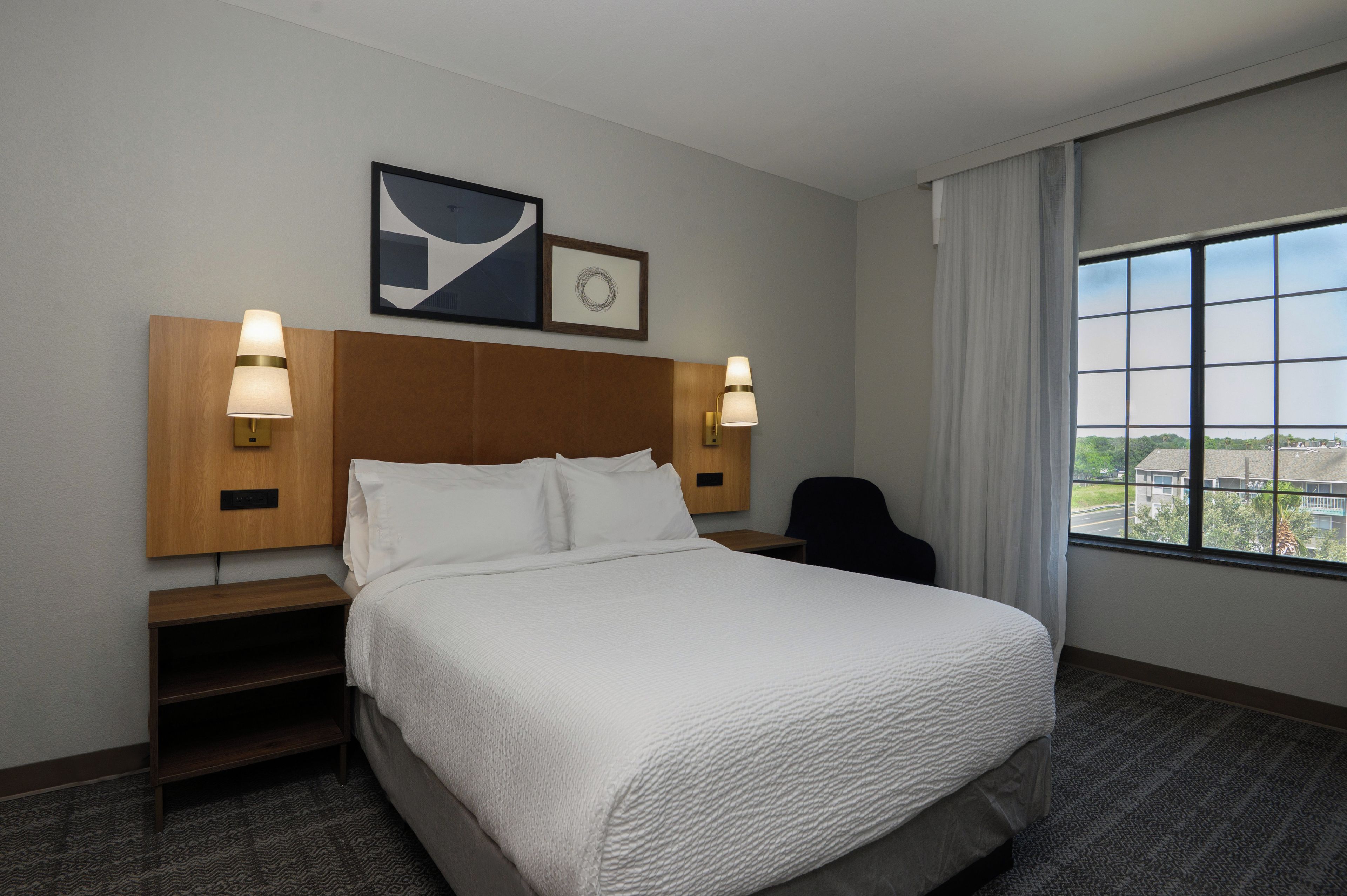 A hotel room with a bed, two bedside lamps, a nightstand, a chair, a window with a view, and two wall art pieces.