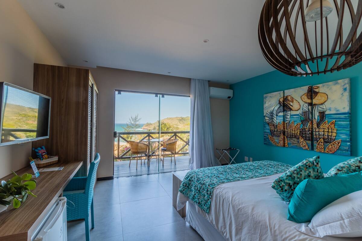 A modern hotel room with a large bed, a TV, a desk with a computer, and a balcony with a view of the sea.