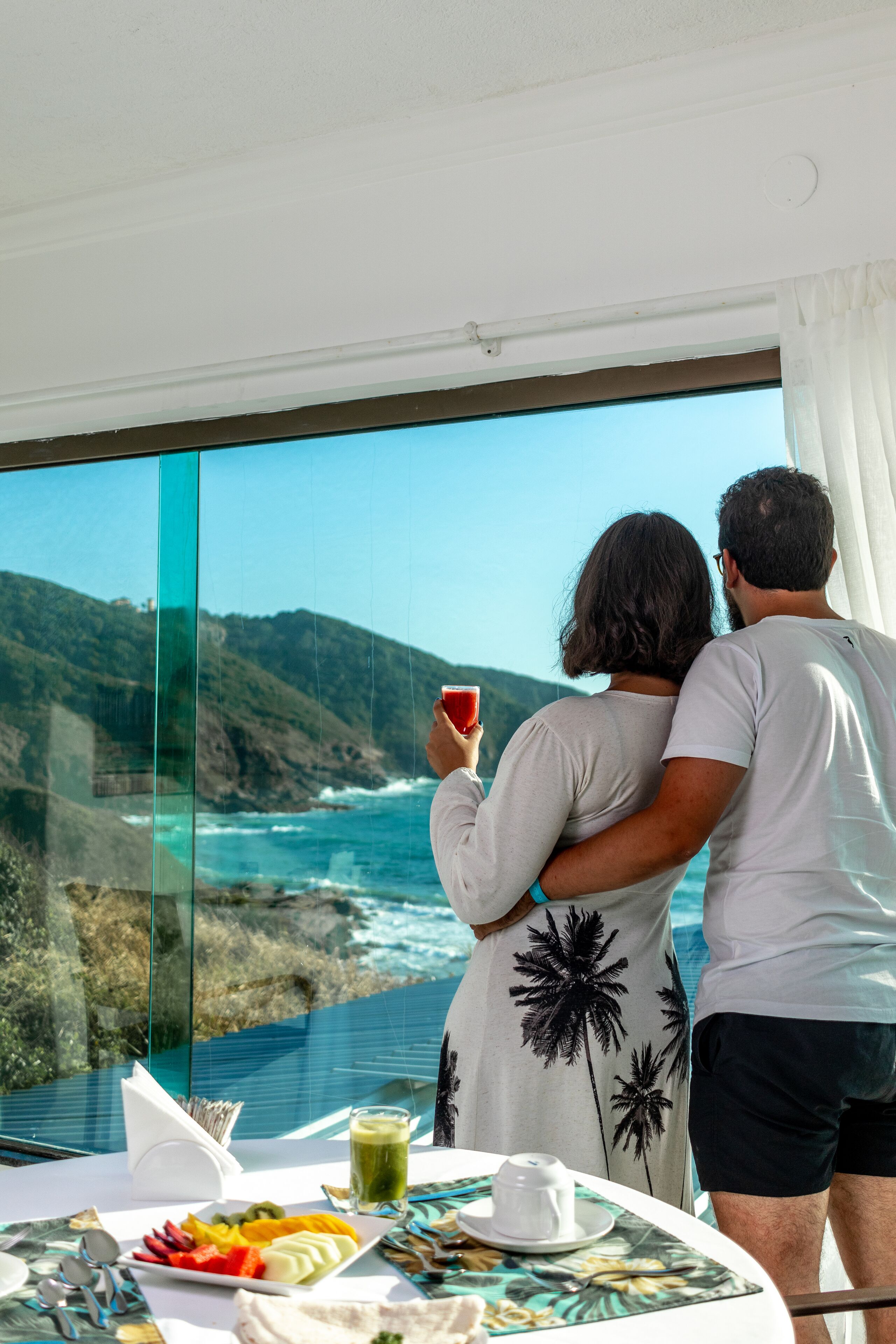 Una pareja admira una vista costera panorámica desde un balcón con una mesa dispuesta para comer.