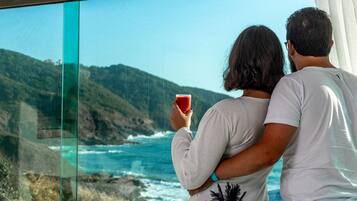 Una pareja admira una vista costera panorámica desde un balcón con una mesa dispuesta para comer.