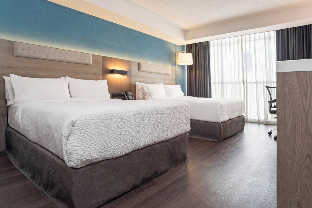 A hotel room with two beds, a wooden headboard, a wall-mounted phone, and a large window with curtains.