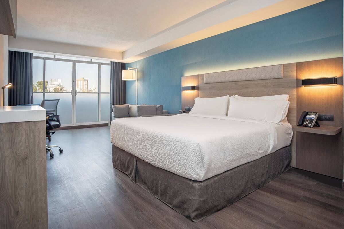 A hotel room with a large bed, a desk, a chair, and a view of the city through large windows.
