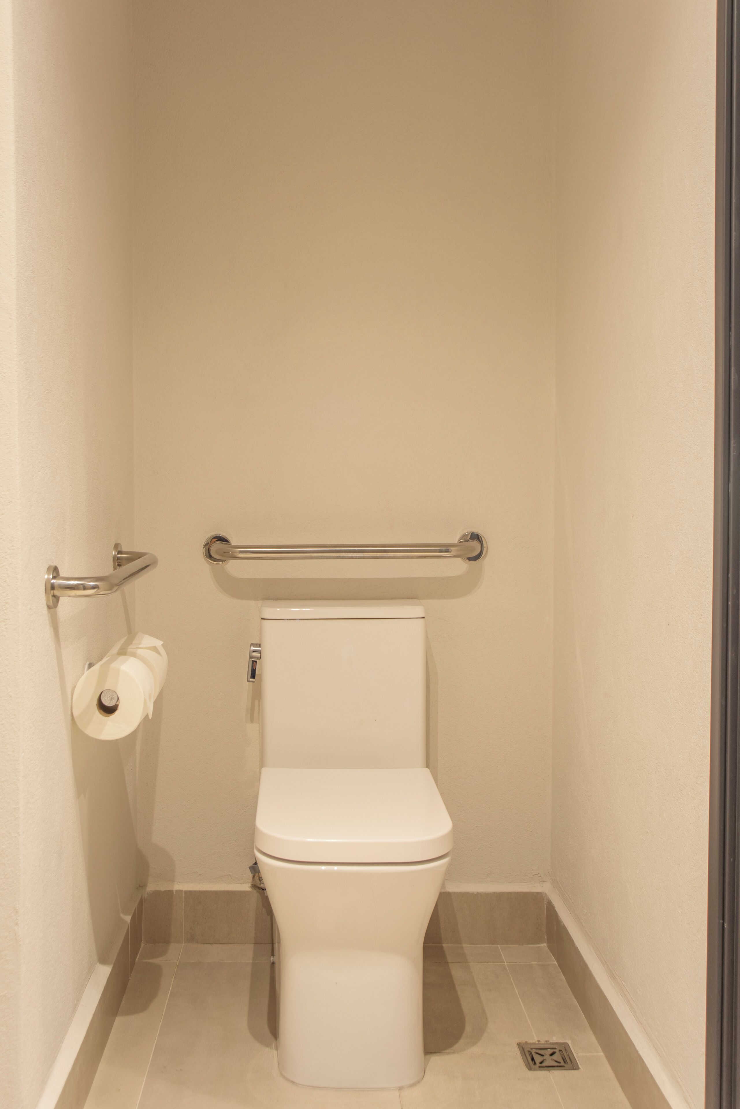 Un baño pequeño, para una sola persona, con inodoro, barra de apoyo y dispensador de papel higiénico.