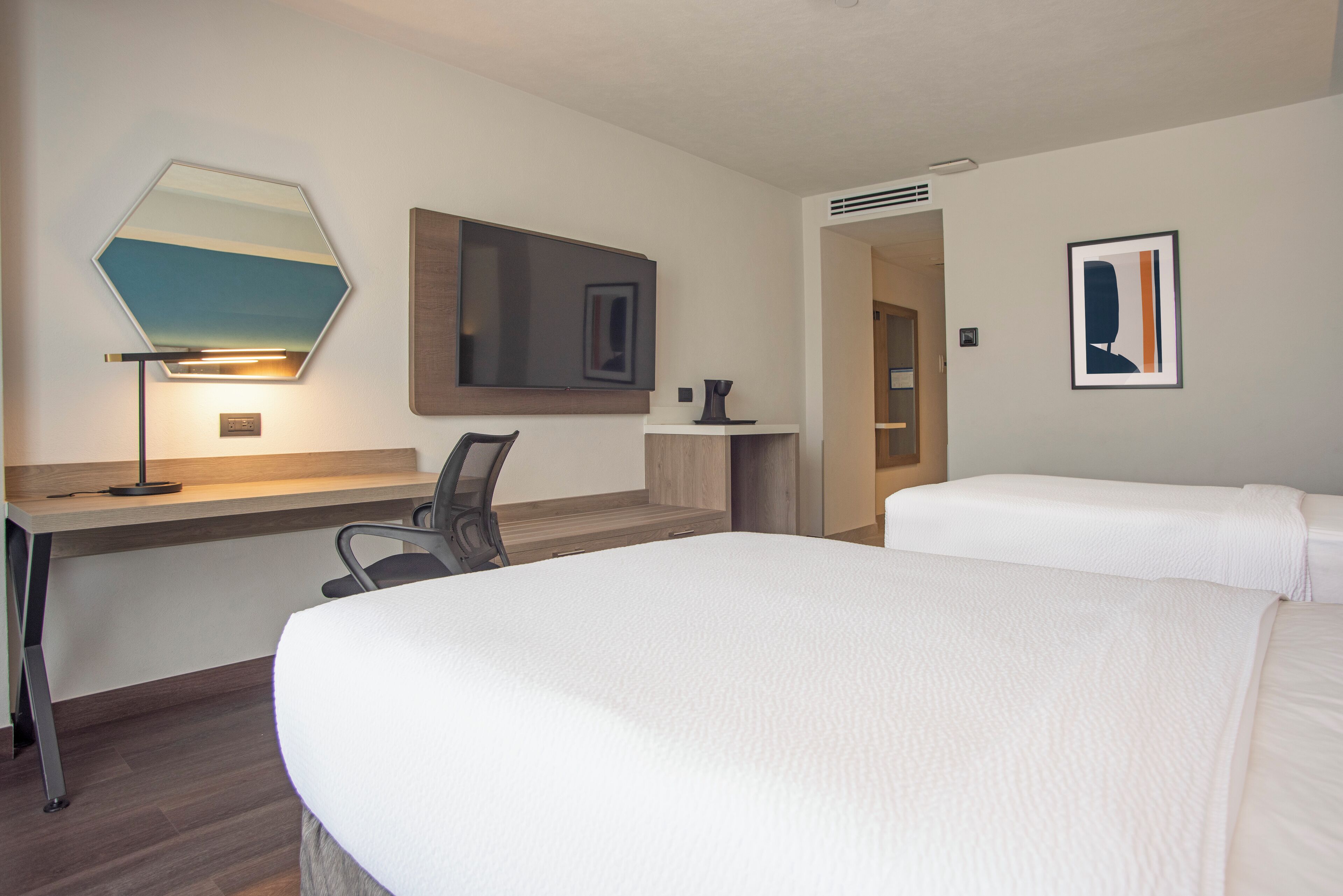 A hotel room with a large bed, a desk with a chair, a mirror, and a framed picture on the wall.