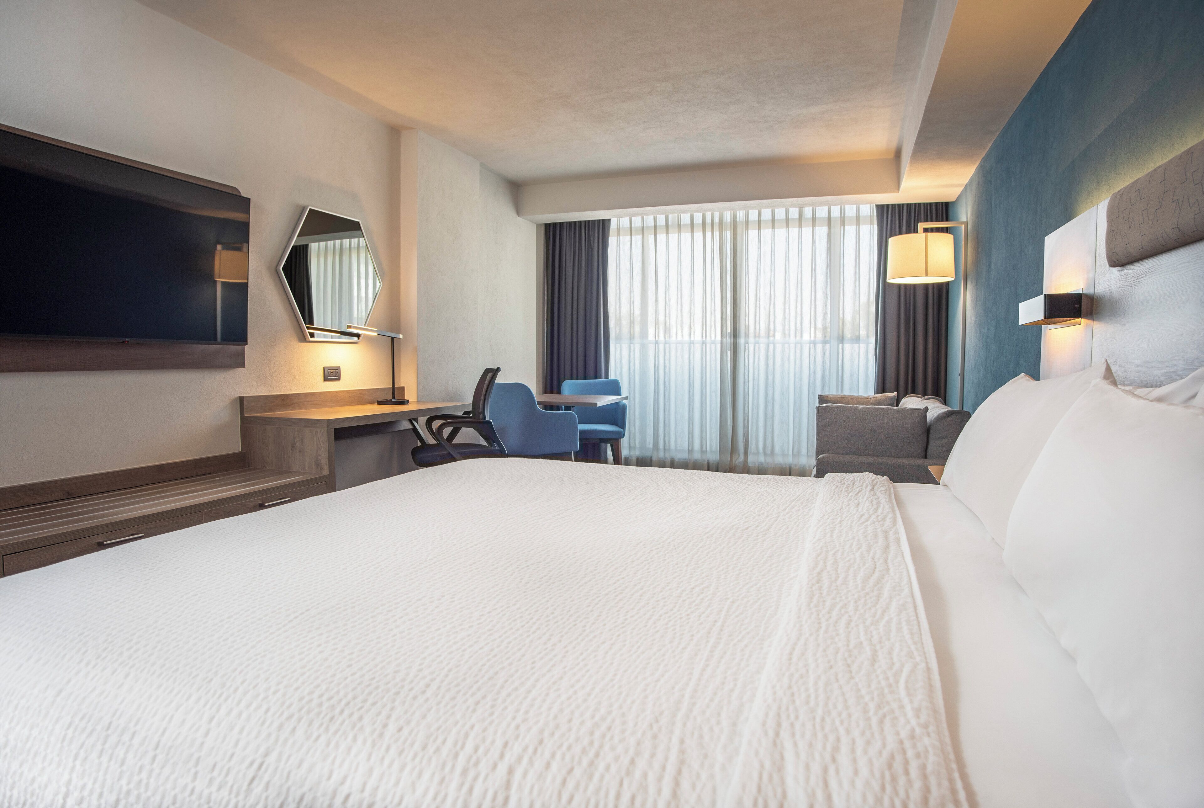 Une chambre d’hôtel avec un grand lit, un bureau avec une chaise, un téléviseur fixé au mur et une fenêtre avec des rideaux.