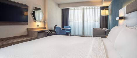 Une chambre d’hôtel avec un grand lit, un bureau avec une chaise, un téléviseur fixé au mur et une fenêtre avec des rideaux.