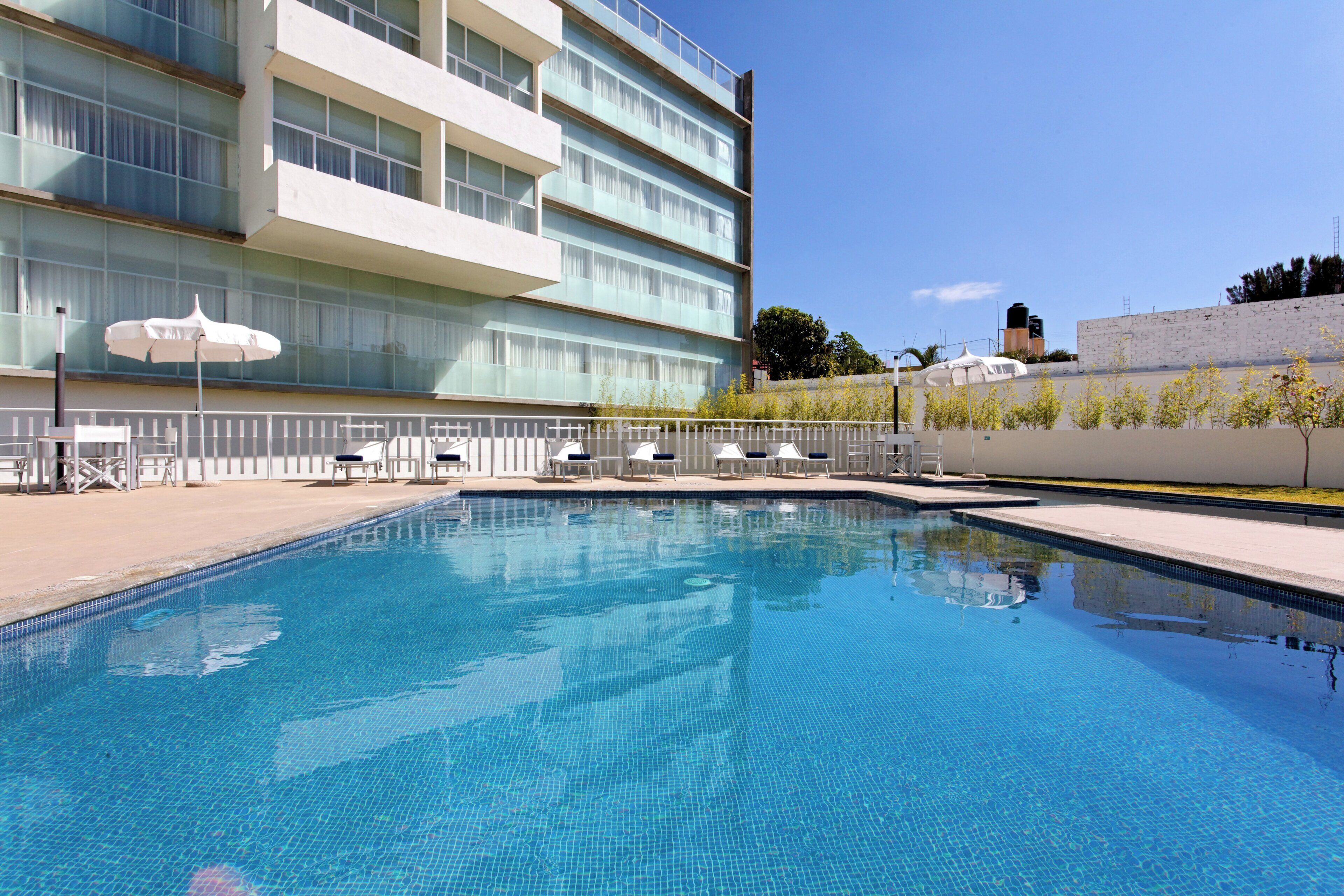 Una piscina con sillones de descanso y sombrillas, rodeada por un edificio de varios pisos y grandes ventanales.