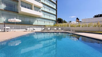 Una piscina con sillones de descanso y sombrillas, rodeada por un edificio de varios pisos y grandes ventanales.