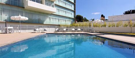 A swimming pool with lounge chairs and umbrellas, surrounded by a building with multiple floors and large windows.