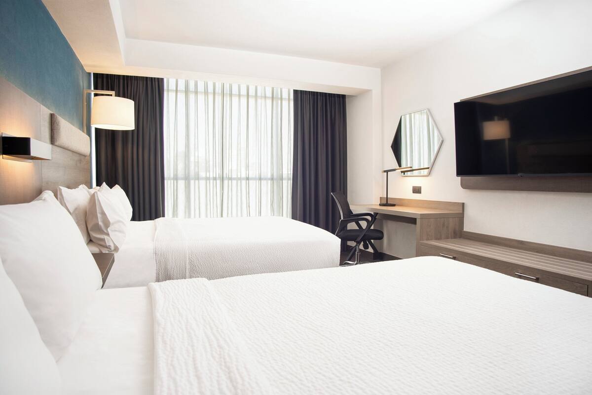 A hotel room with two beds, a desk, a chair, a TV, and a window with curtains.