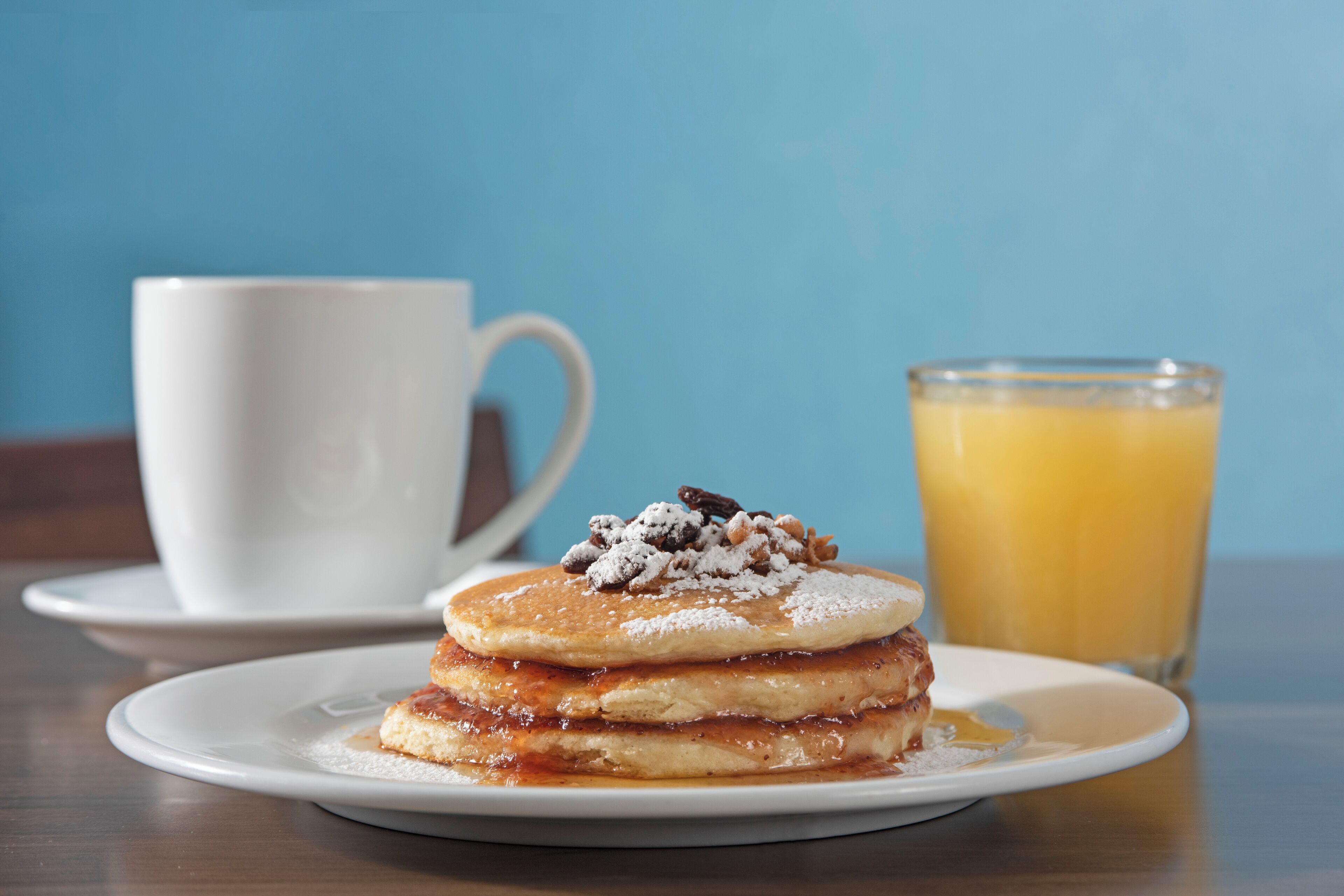 A stack of pancakes with powdered sugar and nuts, a white cup, and a glass of orange juice on a table.