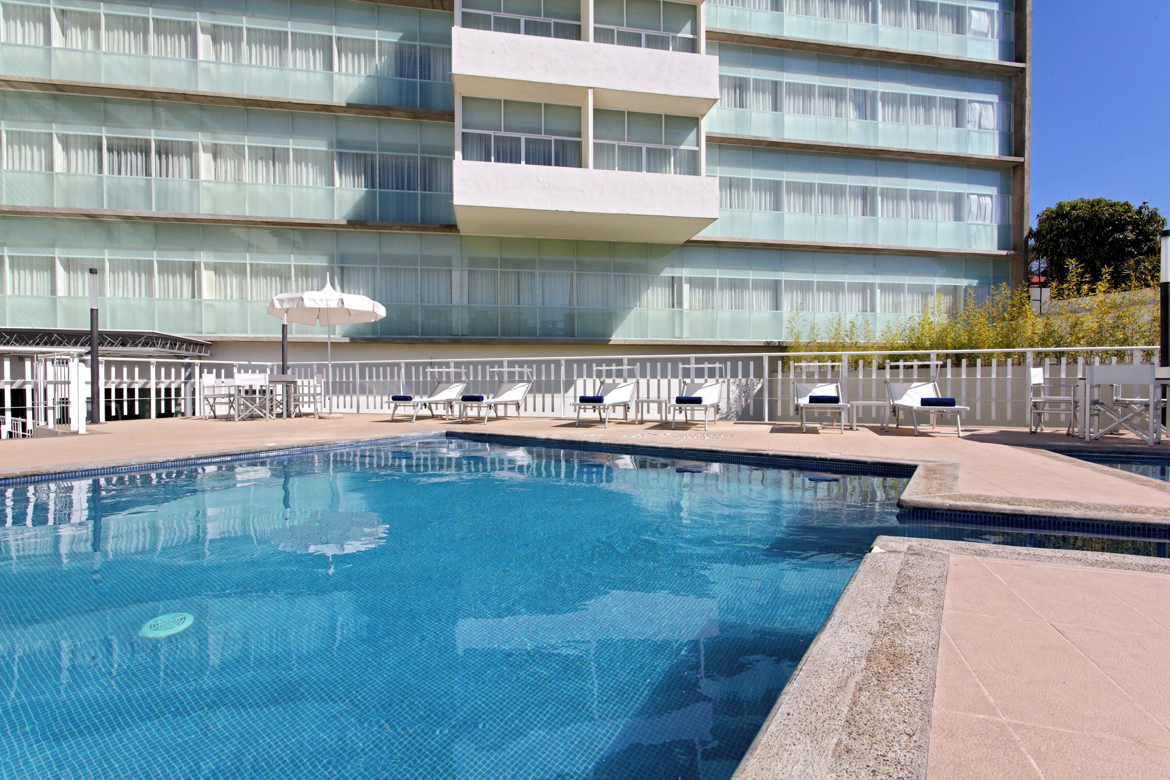 Una piscina con sillones de descanso y sombrillas, rodeada por un edificio de varios pisos y grandes ventanales.