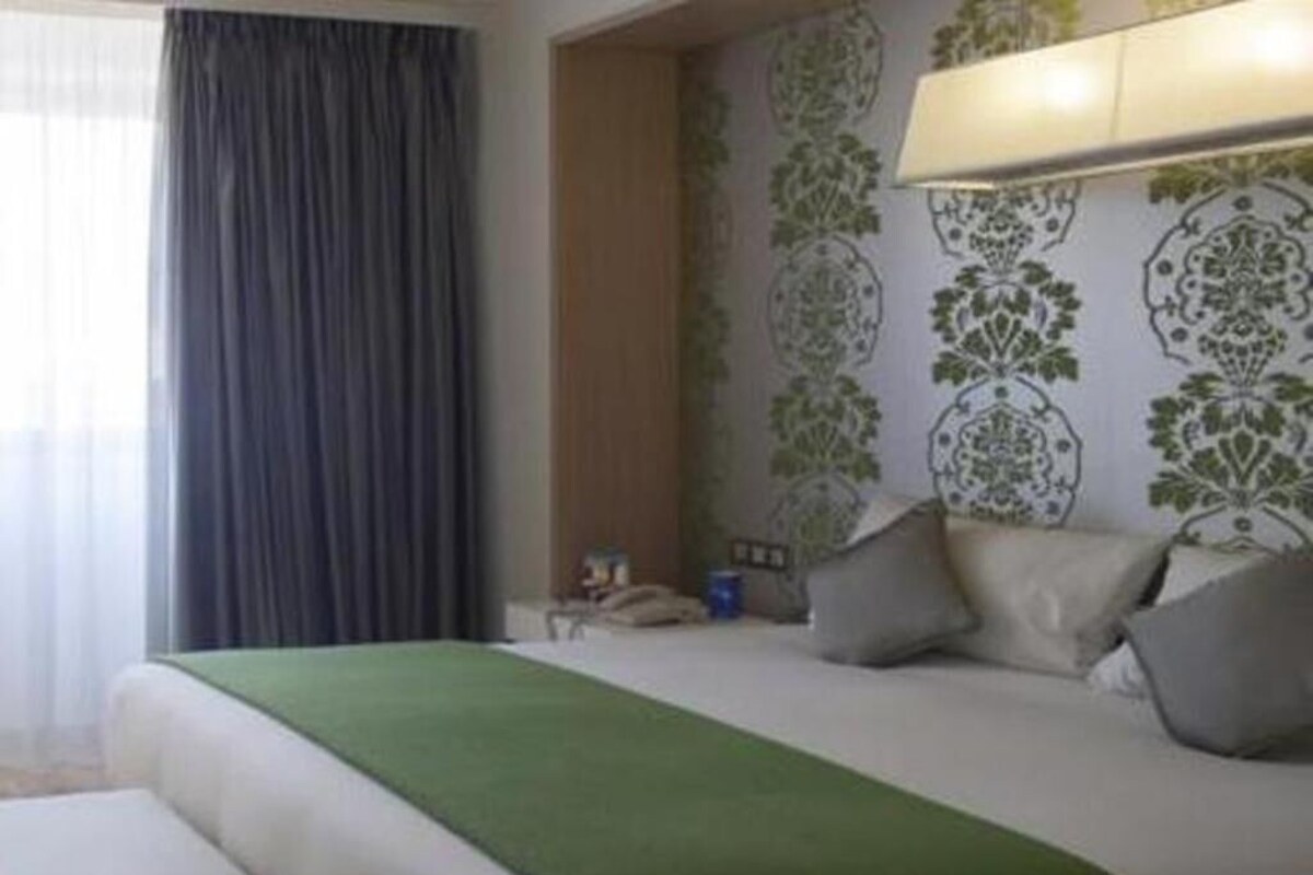 A hotel room with a bed, pillows, a bedside table, and a lamp.