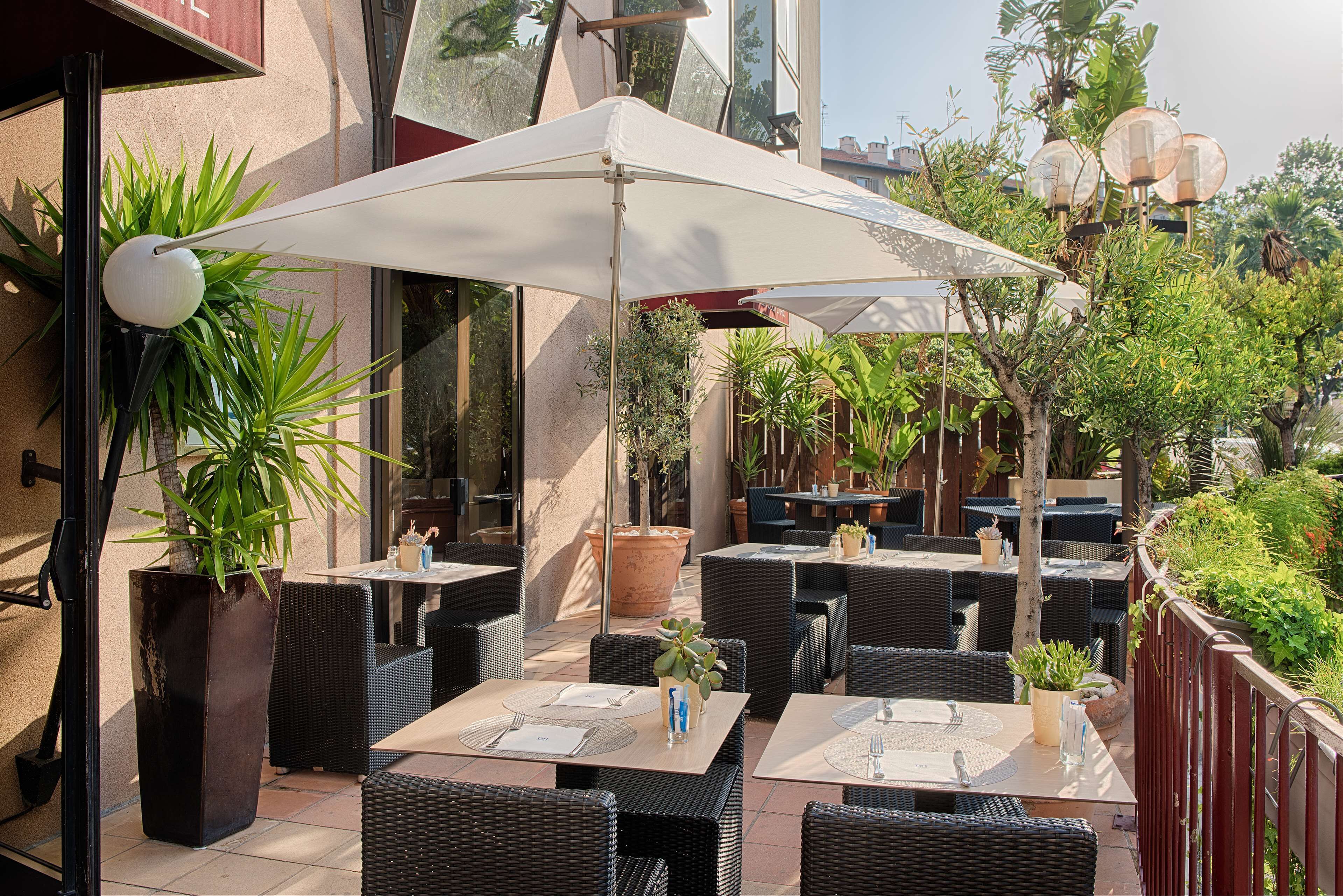 An outdoor patio with tables and chairs, surrounded by plants and a large umbrella.
