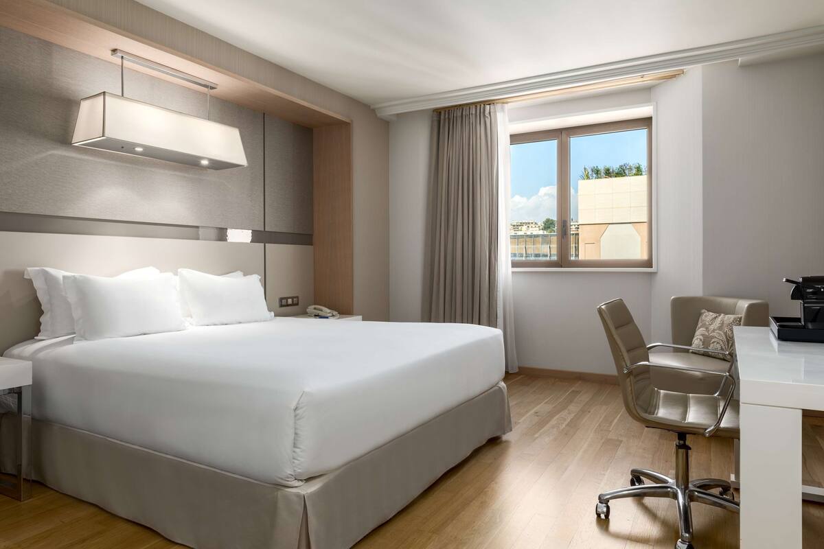 A hotel room with a large bed, a desk, a chair, and a window with curtains.