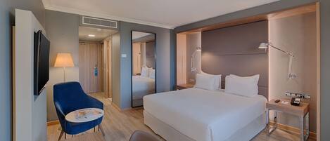 A modern hotel room with a large bed, a desk with a lamp, a chair, and a television on the wall.