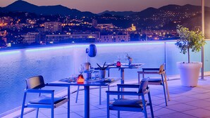 A terrace with tables and chairs, overlooking a cityscape and water at dusk. - NH Nice (Nice)