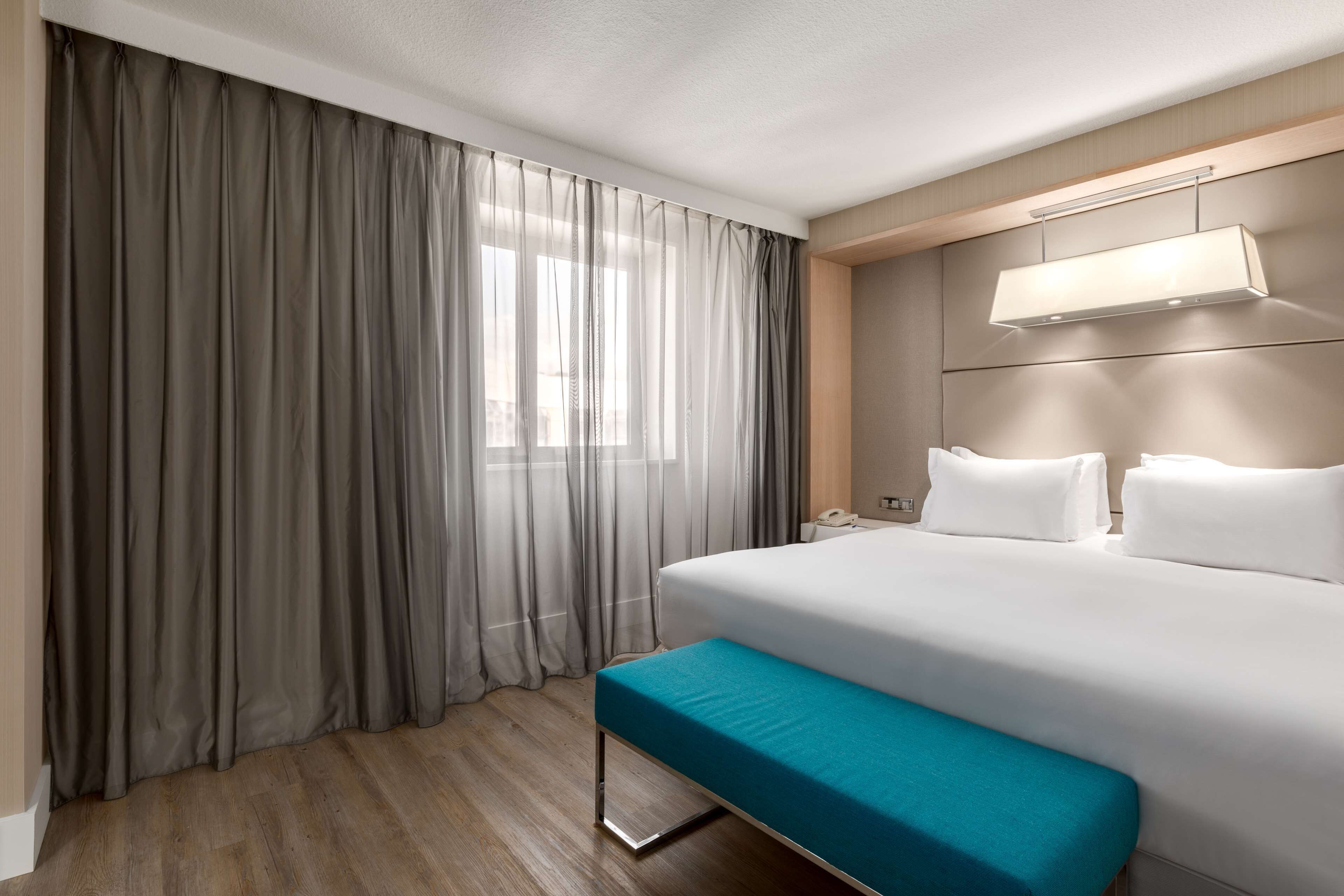 Quarto de hotel com uma cama grande, um banco, mesa de cabeceira, luminária e janela com cortinas.