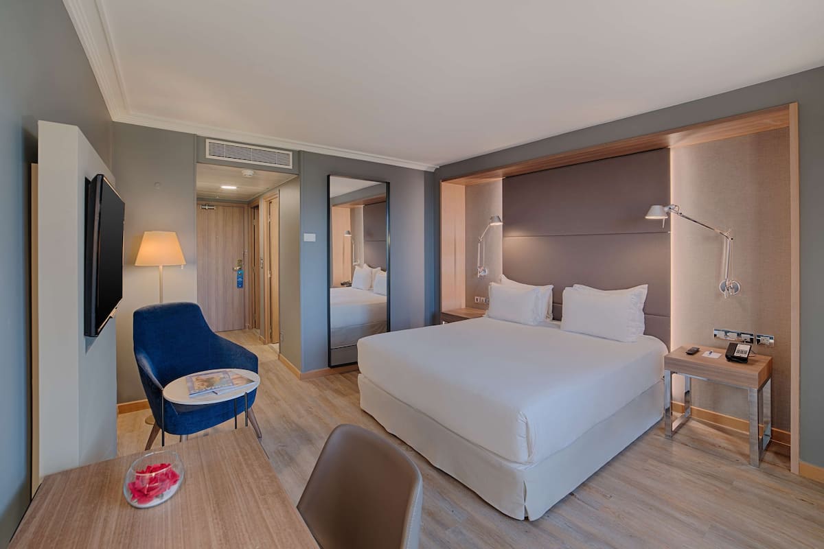 A hotel room with a large bed, a desk with a lamp, a chair, a TV, and a mirror.