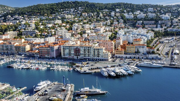 A marina with numerous boats docked, surrounded by a densely packed urban area with various buildings and a hilly backdrop. - NH Nice (Nice)
