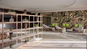 A modern lobby with a wooden shelving unit displaying various plants, a seating area with white chairs and a sofa, and a large wall mural. - NH Nice (Nice)