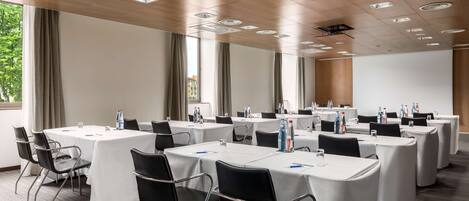 A conference room with multiple tables and chairs, large windows with curtains, and a ceiling with recessed lighting.