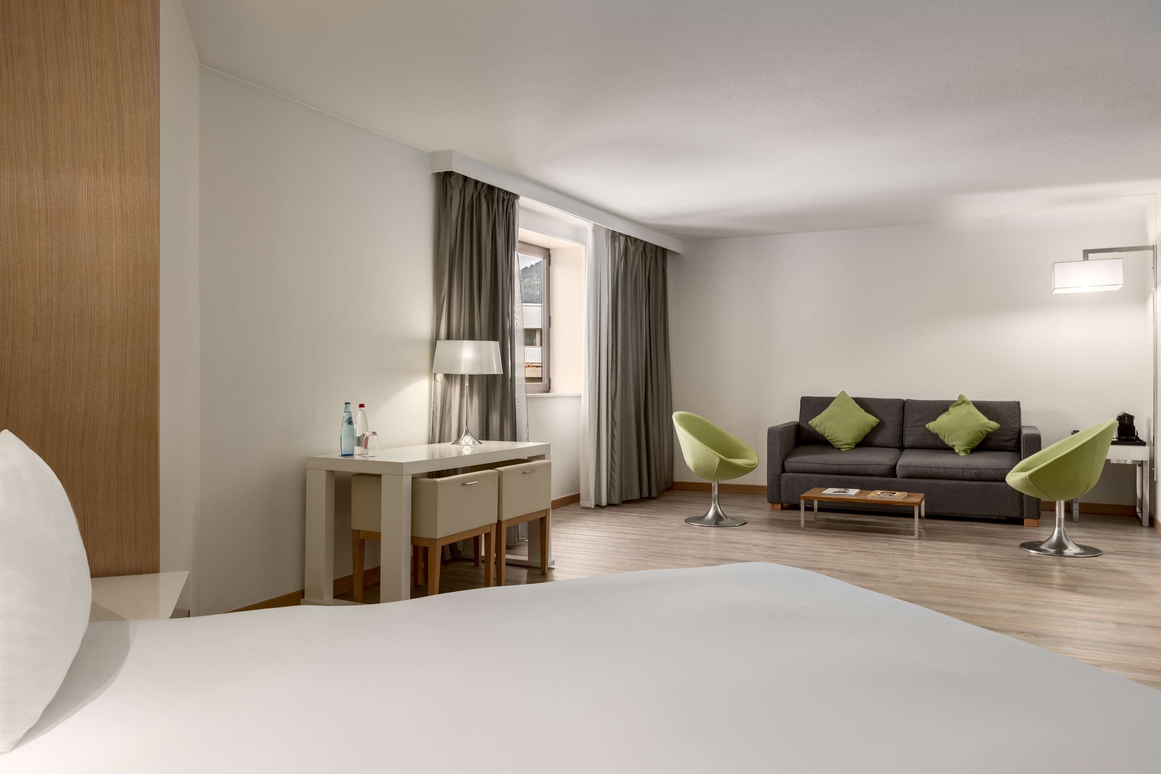 A modern hotel room with a large bed, a desk with a lamp, a sofa with green cushions, and a chair.