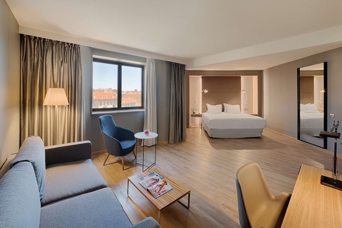 A modern hotel room with a sofa, armchair, small table, and a bed with a nightstand.