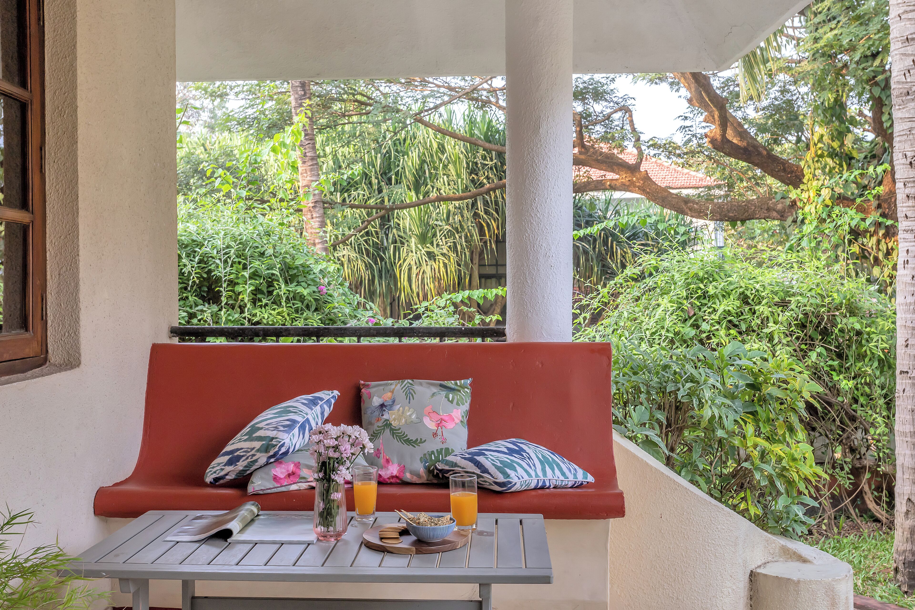 A red bench with floral cushions, a table with a newspaper, a vase with flowers, and two glasses of orange juice.