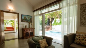 A living room with a sofa, a coffee table, and a view of a swimming pool. - Aguada Anchorage - The Villa Resort (Candolim)