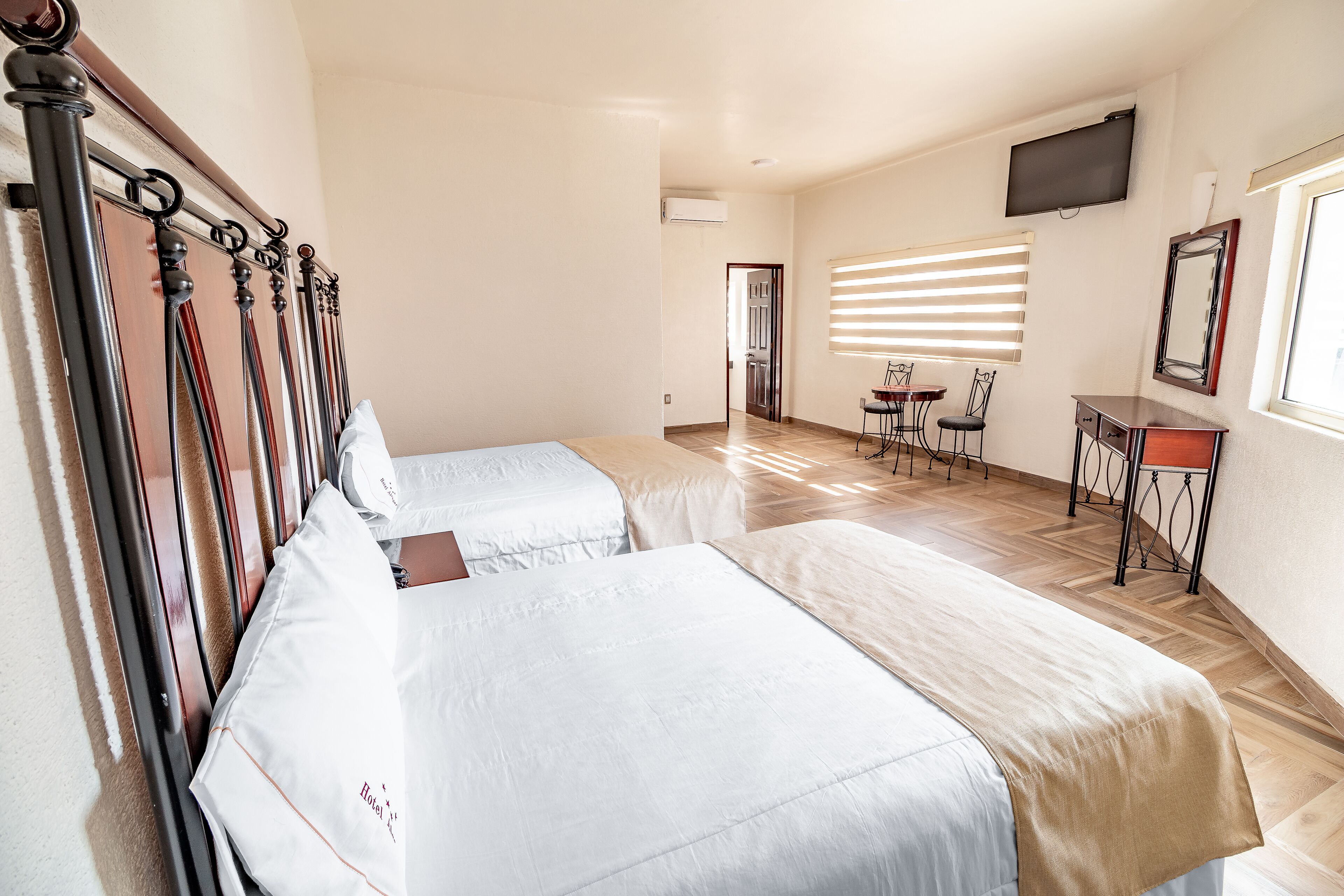 A hotel room with two beds, a TV mounted on the wall, a small table with chairs, and a window with blinds.