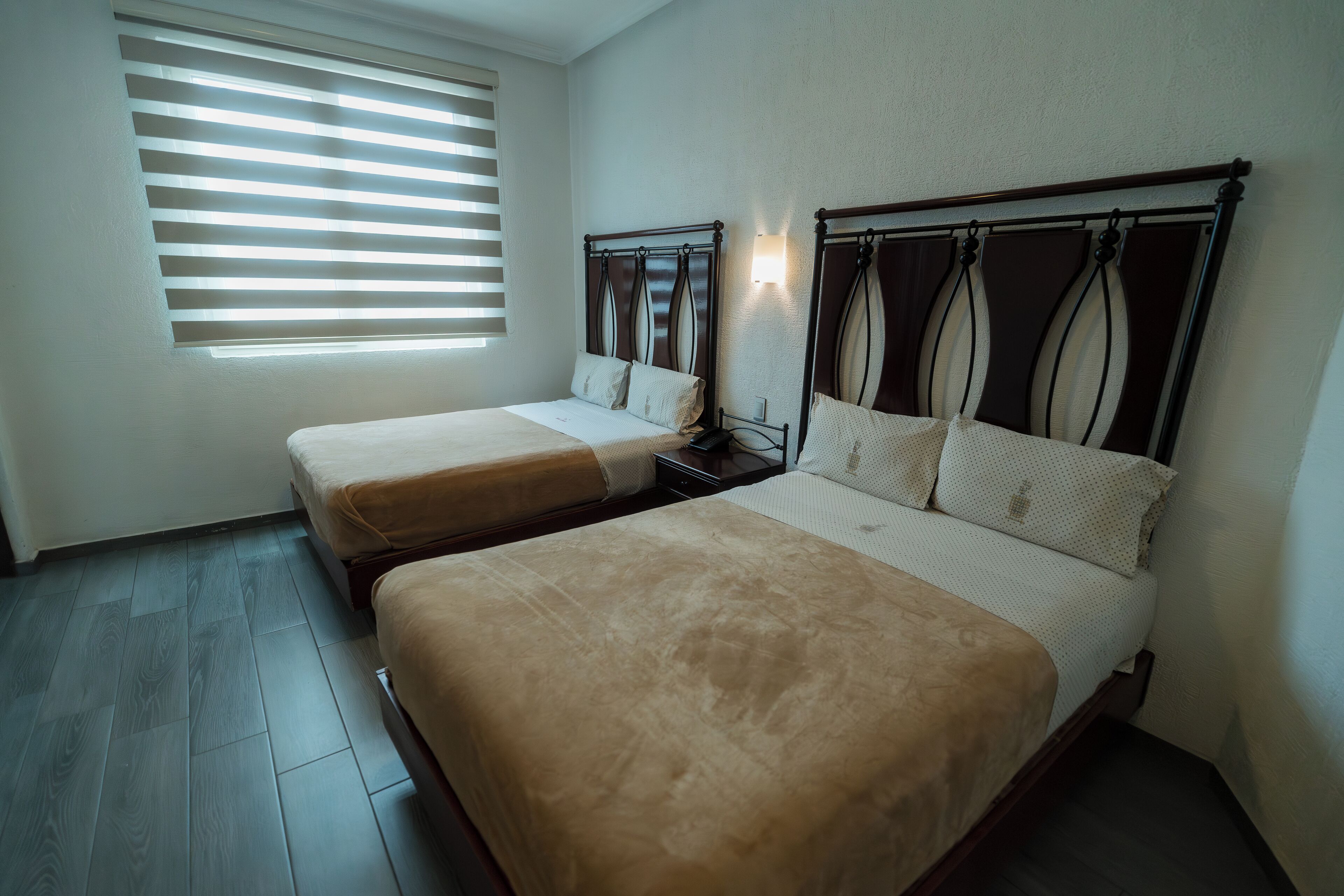 Une chambre d’hôtel avec deux lits, une fenêtre avec des stores, une table de chevet et un luminaire fixé au mur.