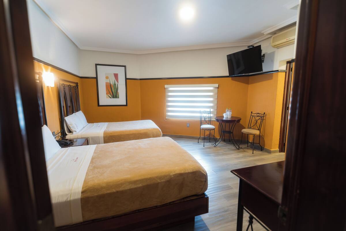 A hotel room with two beds, a small table, a chair, a TV, and a window with blinds.