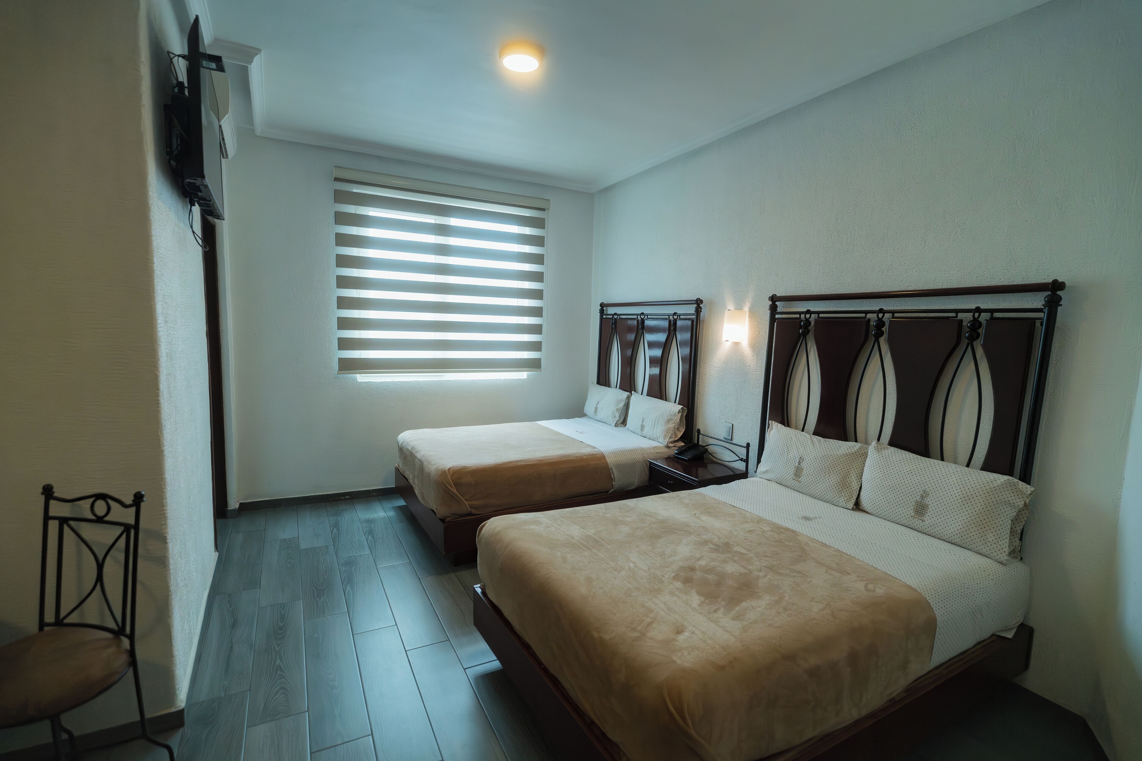 A bedroom with two single beds, a chair, a window with blinds, and a wall-mounted TV.