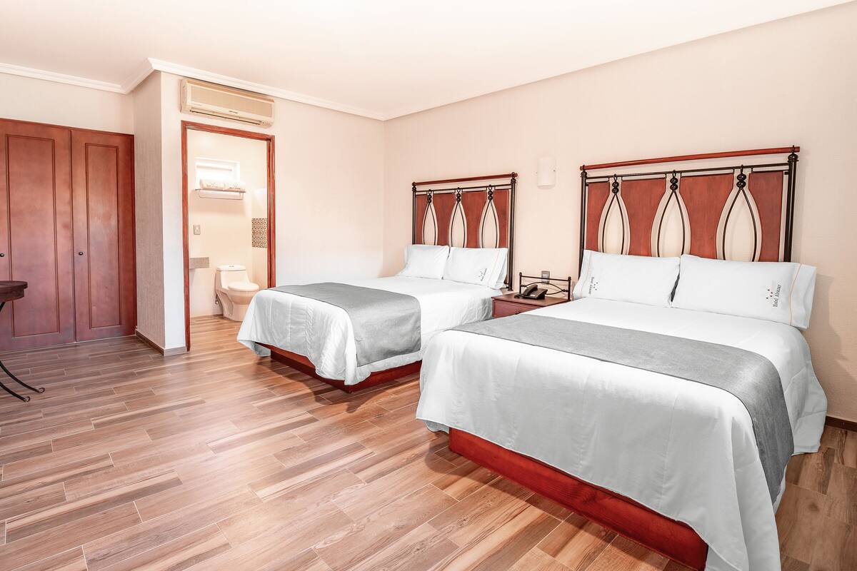 A hotel room with two beds, a wooden floor, a bathroom with a toilet and sink, and a small table with chairs.